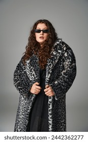 attractive plus size young woman in leopard fur coat and sunglasses standing on grey background