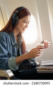 Attractive Portrait Of An Asian Woman Sitting At A Window Seat In Economy Class Listening To Music During A Flight On A Plane, Travel Concept, Vacation, Relaxation