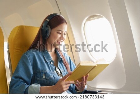 Attractive portrait of an Asian woman sitting in a window seat in economy class reading a book and listening to instrumental music during an airplane flight, travel concept, vacation, relaxation