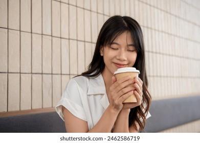 An attractive, positive Asian woman is keeping her eyes closed, smelling the coffee, while holding a takeaway coffee cup and enjoying her coffee in a cafe. coffee break, breakfast, people and drinks - Powered by Shutterstock