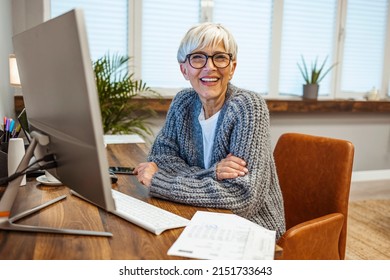 An Attractive Positive Talented Mature Woman Sits In Front Of A Computer And Watches The Bills She Has To Pay, Looking At The Camera With A Happy Smile. The Senior Makes Notes In A Notebook