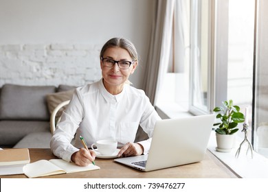 Attractive Positive Talented Mature Woman Writer Sitting In Front Of Laptop And Writing Her New Book About Love, Looking At Camera With Joyfull Smile. Senior Female Teacher Making Notes In Copybook