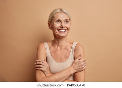 Attractive positive wrinkled fifty years old woman looks gladfully above keeps arms foded has well cared complexion healthy skin white teeth isolated over brown background. Beauty and age concept