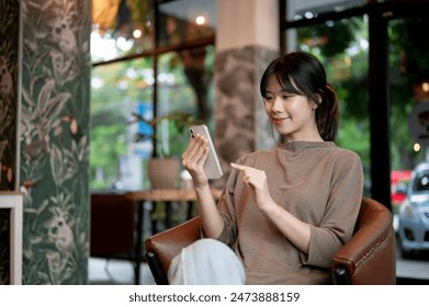 An attractive, positive young Asian woman is using her smartphone on an armchair in a modern coffee shop. chatting, shopping online, mobile app, social media - Powered by Shutterstock