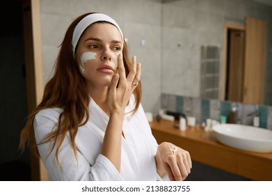 Attractive pregnant red haired woman in white bathrobe standing in the bathroom and taking care of her face skin, applying moisturizer or facial exfoliating scrub, doing smoothing massage movements