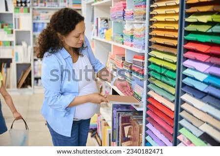 Attractive pregnant woman choosing color paper for design, pastel drawing or watercolor painting, standing by shelves with stationary for creative fine art, in art stationary shop