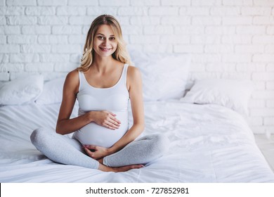 Attractive pregnant woman is sitting in bed and holding her belly. Last months of pregnancy.