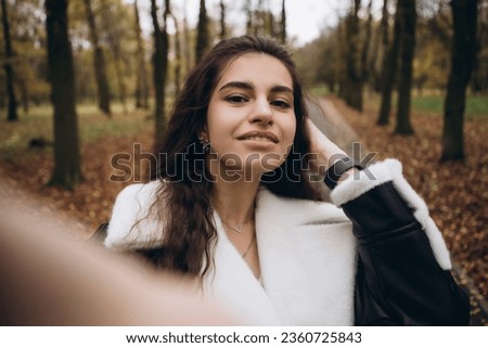 Attractive pretty woman make selfie outdoor, portrait of young lady in warm sunny autumn park season, fall, yellow orange red leaves, dressed leather wool fur jacket smiling having fun 
