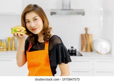 Attractive Pretty Woman Wears Orange Apron Standing Beside Flying Pan In Modern Home Kitchen Smiling And Proud Looking At Tasty Hamburger On Hand That She Learned To Cook For Kid Of Family.