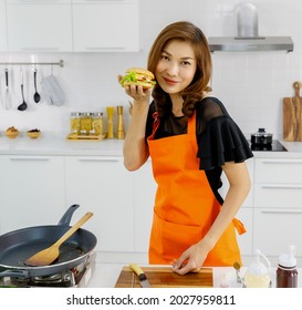 Attractive Pretty Woman Wears Orange Apron Standing Beside Flying Pan In Modern Home Kitchen Smiling And Proud Looking At Tasty Hamburger On Hand That She Learned To Cook For Kid Of Family.