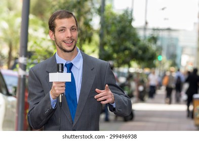 Attractive Professional Male News Reporter Wearing Grey Suit Holding Microphone, Talking To Camera From Urban Setting.