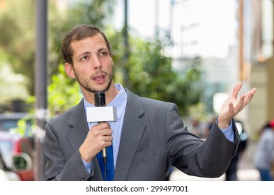 Attractive Professional Male News Reporter Wearing Grey Suit Holding Microphone, Talking To Camera From Urban Setting.