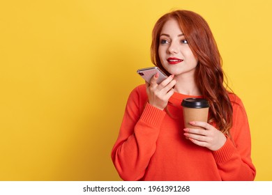 Attractive red haired woman recording voice messages, holding cell phone and coffee to go in hands, looking away, copy space for advertisement, isolated over yellow background. - Powered by Shutterstock