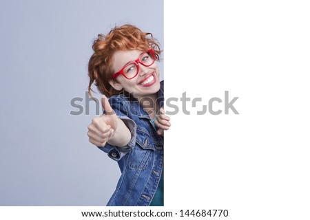 Attractive red-haired girl with red rimless glasses and denim jacket showing thumb up holding large white poster with empty space where you can place your text. Big beautiful eyes. Gray background.