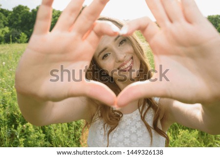 Attractive redhead with natural beauty Caucasian young woman with freckles in a dress looks at the camera, smiles and shows a close-up of a heart shape gesture in nature on a warm summer day.