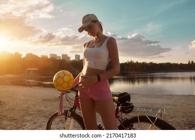 Attractive Redhead Young Woman Riding A Bike On The Beach In Shorts. Plays Volleyball With The Ball. Evening. Sunset. Sand