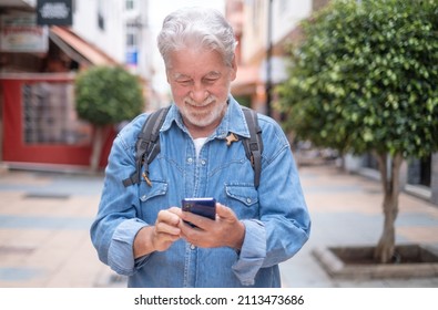 Attractive Relaxed Mature Adult Caucasian Man White- Haired Walking In City Street Holding Cellphone In Hand. Beautiful Senior Man Denim Dressed Enjoys Free Time And Smiles Looking At Smart Phone