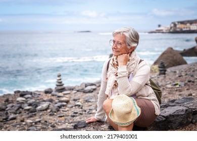 Attractive Relaxed Senior Woman Sitting On A Pebble Beach Looking Away Pensive, Elderly Caucasian Female Enjoying Travel Vacation Retirement