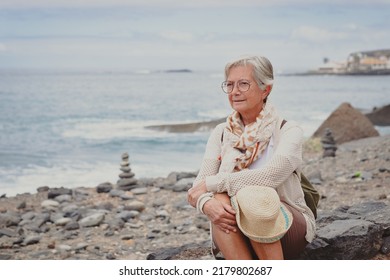 Attractive Relaxed Senior Woman Sitting On A Pebble Beach Looking Away Pensive, Elderly Caucasian Female Enjoying Sea, Travel Vacation Retirement