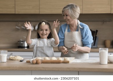 Attractive retired granny teach cute funny granddaughter to cook, prepare buns or cookies, make handmade pastries, happy child throwing dough looks playful white cooking with loving grandma in kitchen - Powered by Shutterstock