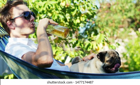 Attractive rich man drinks a beer while resting in hammock with pug dog in the garden on sunset, best friends