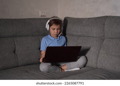 Attractive schoolboy sits on the couch with headphones and a laptop. - Shutterstock ID 2265087791