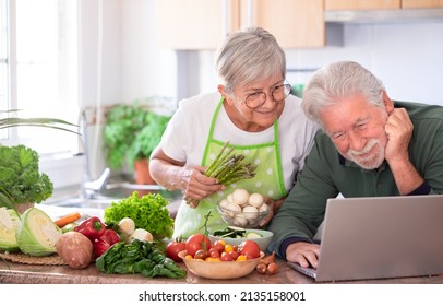 Attractive Senior Couple Preparing Vegetables In Home Kitchen By Trying New Recipes On The Web From Laptop. Smiling Caucasian Seniors Enjoying Healthy Nutrition