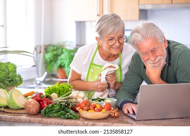 Attractive Senior Couple Preparing Vegetables In Home Kitchen By Trying New Recipes On The Web From Laptop. Smiling Caucasian Seniors Enjoying Healthy Nutrition