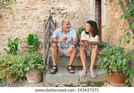 Attractive senior couple sitting on the steps of a luxury holiday villa with stone walls during a summer holiday, drinking wine and having a conversation. Mature people vacation outdoors lifestyle.