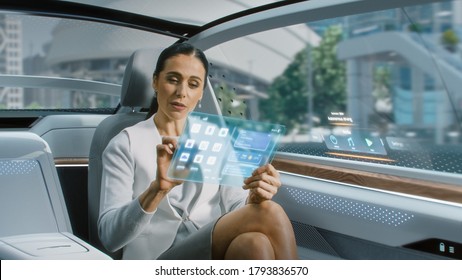 Attractive Senior Female Reading News on a Futuristic Transparent Tablet Computer with Augmented Reality Interface while Sitting on a Backseat of Autonomous Car. Self-Driving Van Rides on Public Road.