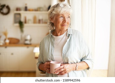 Attractive senior gray haired woman wearing round spectacles on her head having calm peaceful morning standing by window with mug, drinking coffee. Elderly female enjoying herbal tea indoors