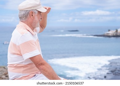 Attractive Senior Man With Beard And Hat Sitting Outdoors At Sea Looking At Horizon Enjoying Freedom And Vacation. Active Caucasian Elderly Man Relaxing On A Sunny Day Close To The Beach