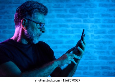 Attractive Senior Man In Eyeglasses Talking On Smartphone. Portrait Of Mature Man Talking On Phone. Portrait Of Man Stands Against A Brick Wall And Gel Light With Mobile Phone