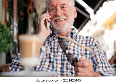 Attractive senior man sitting outdoor at a cafe table talking on mobile phone while enjoying a coffee and milk drink - caucasian elderly man with glasses in hand relaxed in retirement or vacation - Shutterstock ID 2231420145
