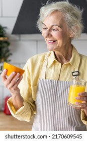 Attractive Senior Woman Drinking Freshly Squeezed Orange Juice, Looking Happy And Healthy. Concept Of Active Lifestyle Of Mature People, Cooking Organic Food. Mature Female At Home At Her Kitchen