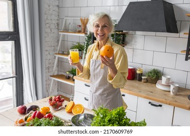 Attractive Senior Woman Drinking Freshly Squeezed Orange Juice, Looking Happy And Healthy. Concept Of Active Lifestyle Of Mature People, Cooking Organic Food. Mature Female At Home At Her Kitchen