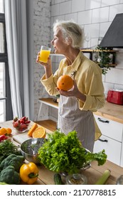 Attractive Senior Woman Drinking Freshly Squeezed Orange Juice, Looking Happy And Healthy. Concept Of Active Lifestyle Of Mature People, Cooking Organic Food. Mature Female At Home At Her Kitchen