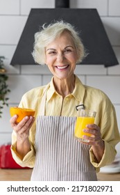 Attractive Senior Woman Drinking Freshly Squeezed Orange Juice, Looking Happy And Healthy. Concept Of Active Lifestyle Of Mature People, Cooking Organic Food. Mature Female At Home At Her Kitchen