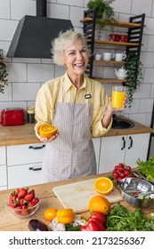 Attractive Senior Woman Drinking Freshly Squeezed Orange Juice, Looking Happy And Healthy. Concept Of Active Lifestyle Of Mature People, Cooking Organic Food. Mature Female At Home At Her Kitchen