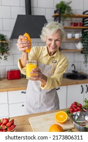 Attractive Senior Woman Drinking Freshly Squeezed Orange Juice, Looking Happy And Healthy. Concept Of Active Lifestyle Of Mature People, Cooking Organic Food. Mature Female At Home At Her Kitchen