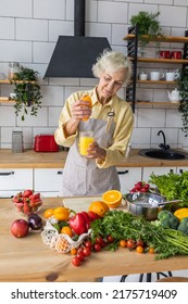 Attractive Senior Woman Drinking Freshly Squeezed Orange Juice, Looking Happy And Healthy. Concept Of Active Lifestyle Of Mature People, Cooking Organic Food. Mature Female At Home At Her Kitchen