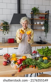 Attractive Senior Woman Drinking Freshly Squeezed Orange Juice, Looking Happy And Healthy. Concept Of Active Lifestyle Of Mature People, Cooking Organic Food. Mature Female At Home At Her Kitchen