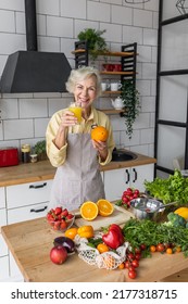 Attractive Senior Woman Drinking Freshly Squeezed Orange Juice, Looking Happy And Healthy. Concept Of Active Lifestyle Of Mature People, Cooking Organic Food. Mature Female At Home At Her Kitchen