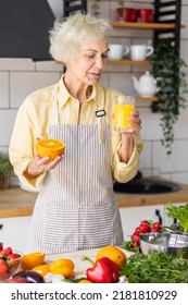 Attractive Senior Woman Drinking Freshly Squeezed Orange Juice, Looking Happy And Healthy. Concept Of Active Lifestyle Of Mature People, Cooking Organic Food. Mature Female At Home At Her Kitchen