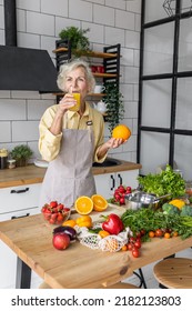 Attractive Senior Woman Drinking Freshly Squeezed Orange Juice, Looking Happy And Healthy. Concept Of Active Lifestyle Of Mature People, Cooking Organic Food. Mature Female At Home At Her Kitchen