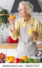 Attractive Senior Woman Drinking Freshly Squeezed Orange Juice, Looking Happy And Healthy. Concept Of Active Lifestyle Of Mature People, Cooking Organic Food. Mature Female At Home At Her Kitchen