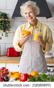 Attractive Senior Woman Drinking Freshly Squeezed Orange Juice, Looking Happy And Healthy. Concept Of Active Lifestyle Of Mature People, Cooking Organic Food. Mature Female At Home At Her Kitchen