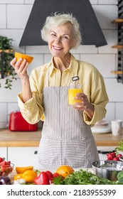 Attractive Senior Woman Drinking Freshly Squeezed Orange Juice, Looking Happy And Healthy. Concept Of Active Lifestyle Of Mature People, Cooking Organic Food. Mature Female At Home At Her Kitchen