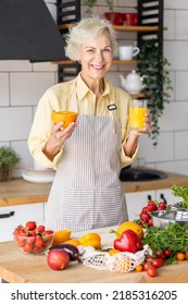 Attractive Senior Woman Drinking Freshly Squeezed Orange Juice, Looking Happy And Healthy. Concept Of Active Lifestyle Of Mature People, Cooking Organic Food. Mature Female At Home At Her Kitchen