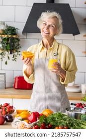 Attractive Senior Woman Drinking Freshly Squeezed Orange Juice, Looking Happy And Healthy. Concept Of Active Lifestyle Of Mature People, Cooking Organic Food. Mature Female At Home At Her Kitchen